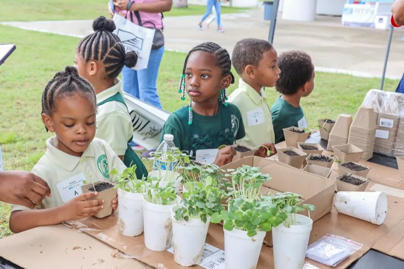 Elementary students in school polos potting seedlings into peat cups at an outdoor RTPark STEM planting station.