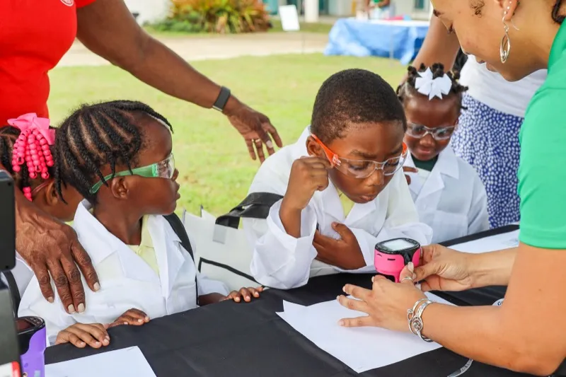 Two students in lab coats and safety goggles examine a sample with a digital microscope while a facilitator guides them.