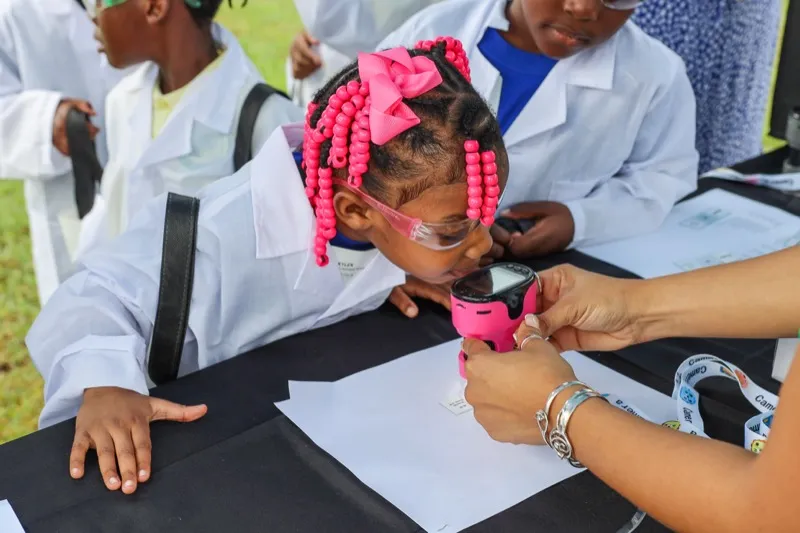 A young student in a lab coat and safety goggles peers into a digital microscope at an outdoor science station.
