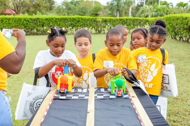 A group of students in matching yellow shirts race handmade toy cars down a wooden ramp on the RTPark lawn.
