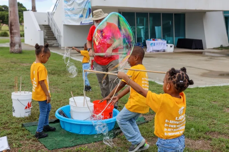 Young students in yellow shirts dip wands into a tray of soapy water to make giant bubbles with a facilitator outdoors.