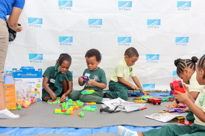 Elementary students sit on a mat building with colorful interlocking blocks in front of an RTPark step-and-repeat backdrop.