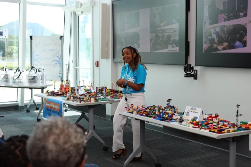 A facilitator in a blue STEM Camp shirt presents in front of tables filled with student-built LEGO city models.
