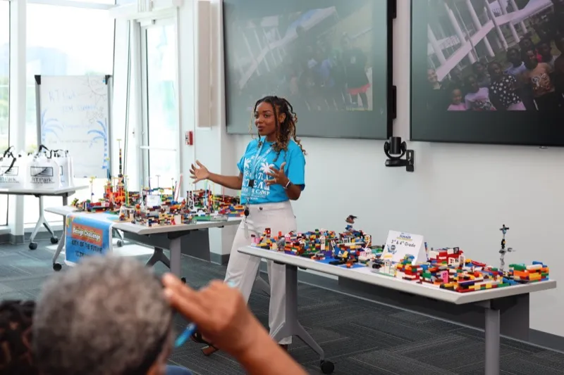 A facilitator addresses the audience while gesturing toward LEGO city models displayed on tables at the STEM Camp showcase.