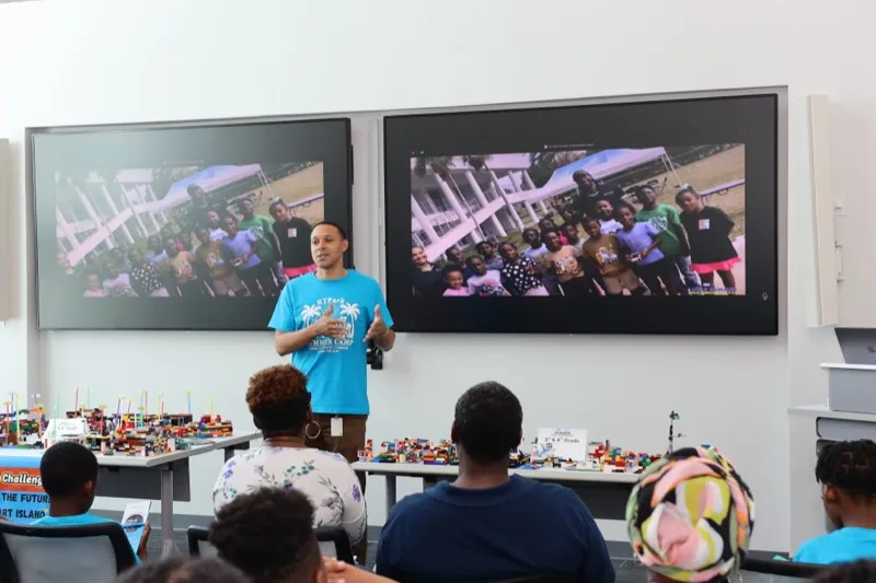 A presenter at the front of a STEM Summer Camp showcase speaks to the audience as camp photos play on the screens behind him.