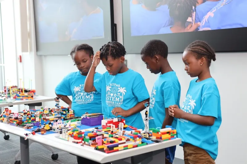 Four campers in blue STEM Summer Camp shirts inspect their finished LEGO city build on a long display table.