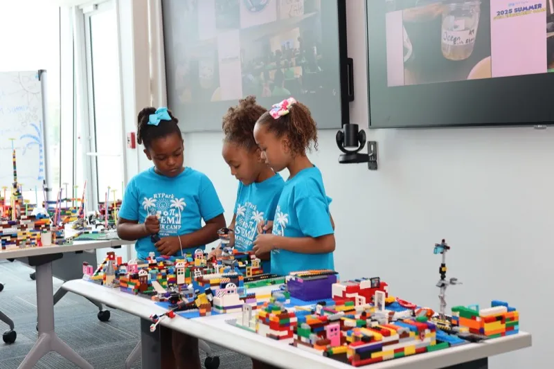 Three young campers in blue STEM Summer Camp shirts add finishing touches to a colorful LEGO cityscape.