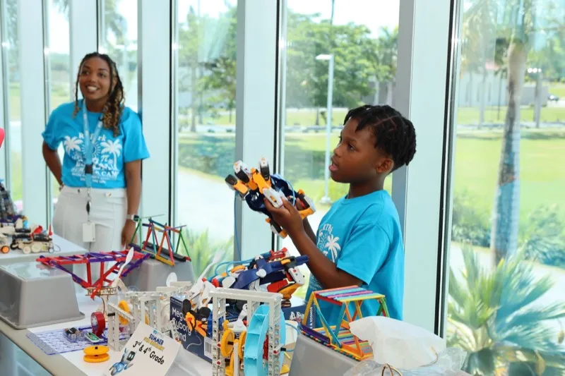 A camper holds up a robotics model he built while a facilitator smiles nearby, with K'NEX projects on the table in front of them.
