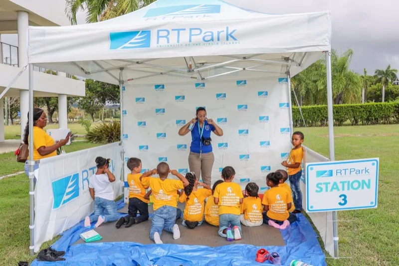 Children seated under an RTPark tent at Station 3 listen to a facilitator during an outdoor STEM rotation.