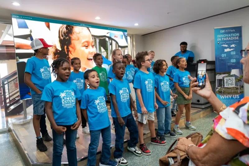 A group of campers in blue STEM Summer Camp shirts sing together in front of a large screen showing camp footage at a closing event.