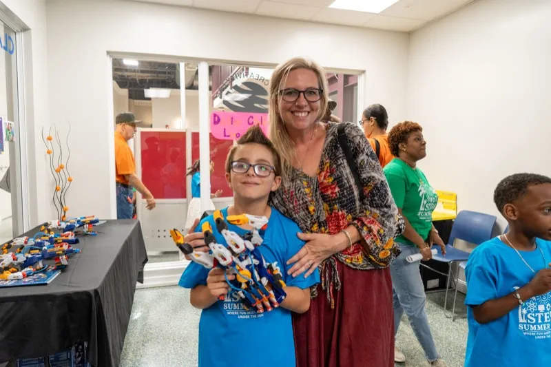 A camper proudly holds up a robotics model he built while posing with his teacher in a STEM Summer Camp classroom.